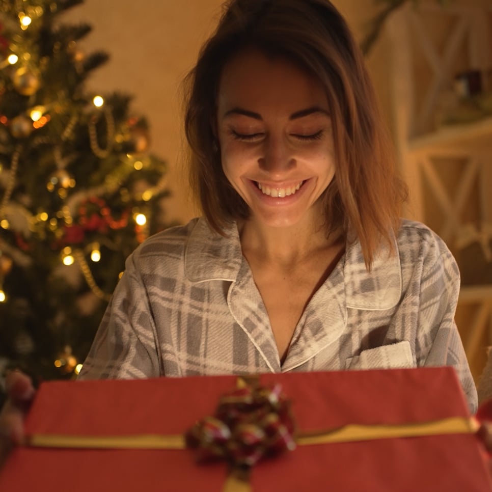 En kvinna öppnar en julklapp framför en julgran.
