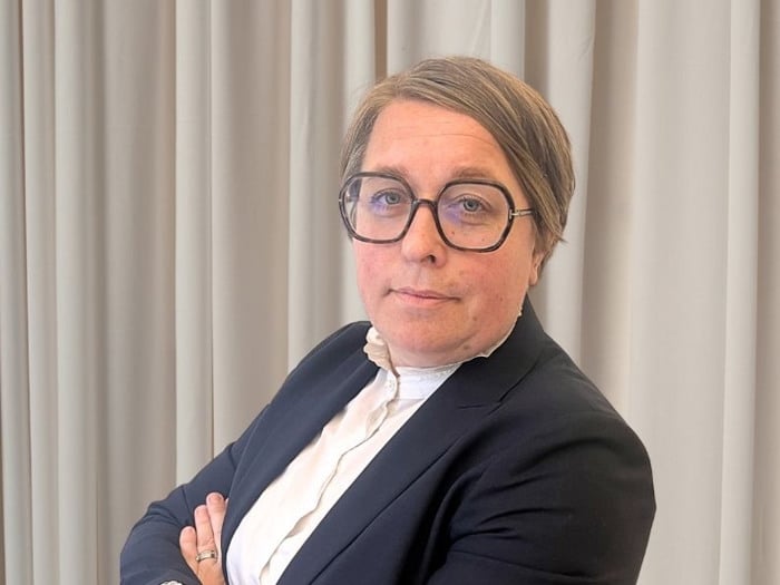 En kvinna i kostym och glasögon som står med armarna i kors.