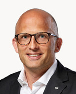 En man i kostym och glasögon ler mot kameran.