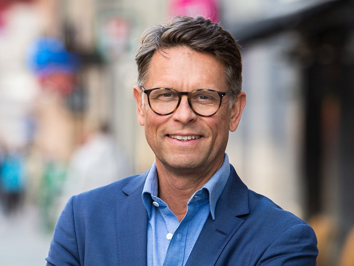 En man i blå kostym och glasögon står på en gata i en stad.